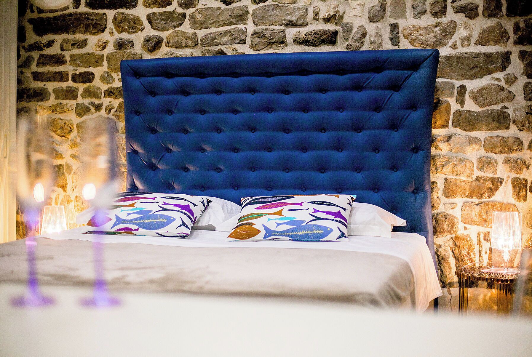 Un lit avec une tête de lit rembourrée de tissu bleu, une literie blanche et deux coussins à motifs.