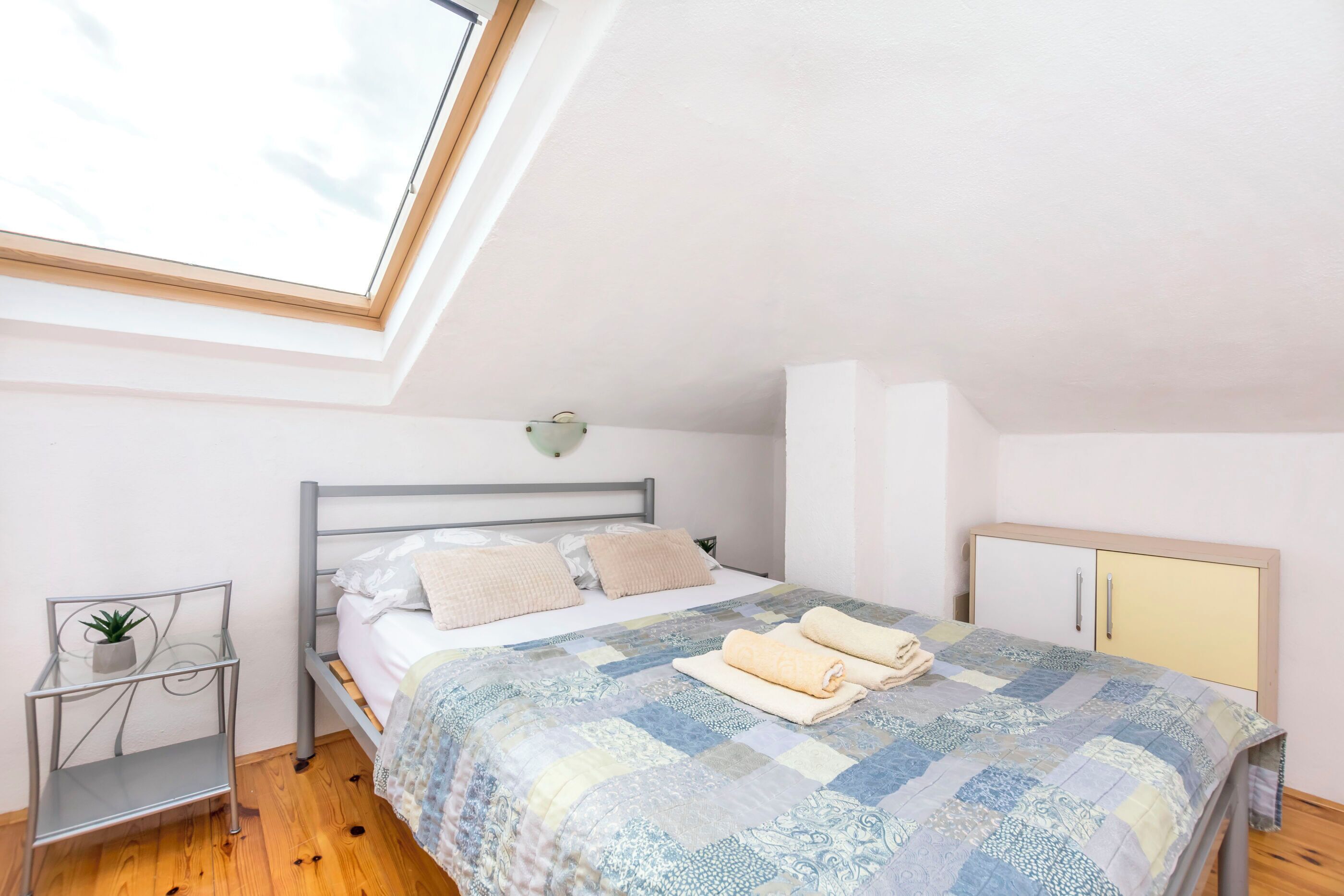 A bedroom with a bed, a bedside table with a plant, a wardrobe, and a skylight.