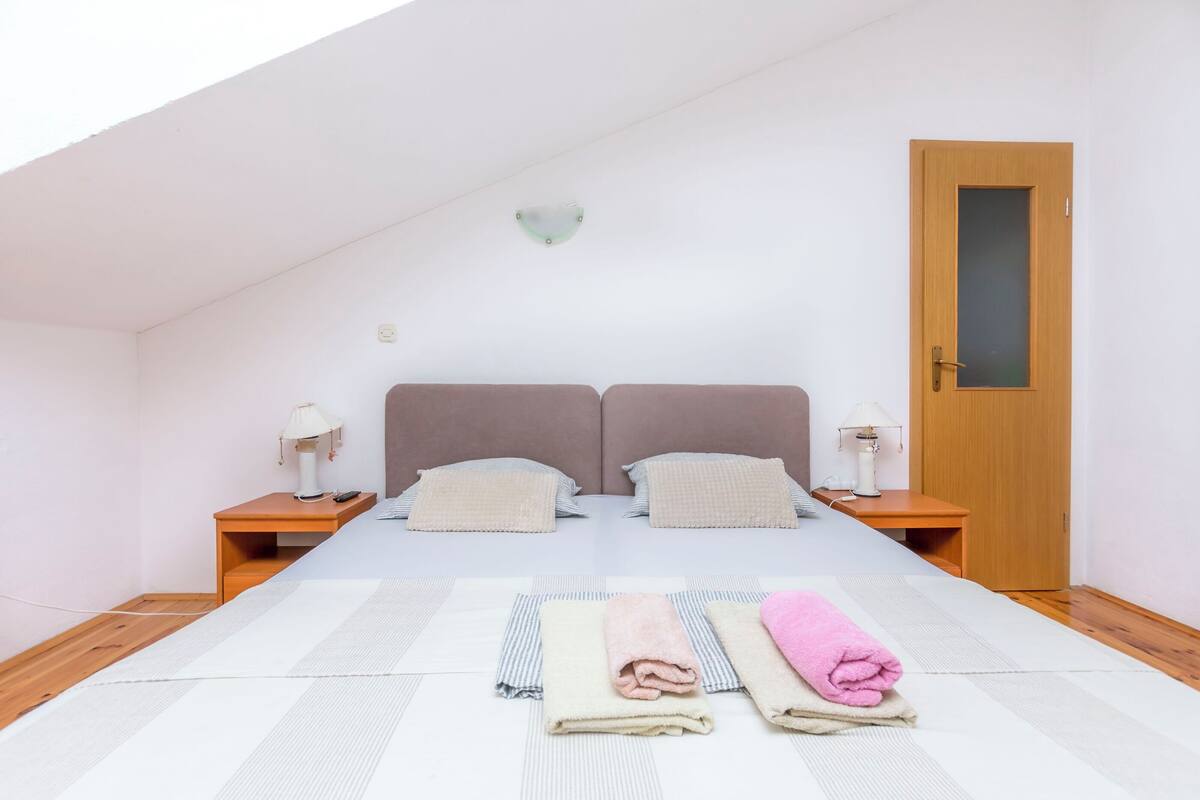 A bedroom with a sloped ceiling, two beds with headboards, bedside tables with lamps, a wooden door, and a view of the sky.