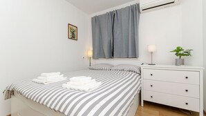 A neatly made bed with striped bedding, a bedside lamp, a small plant, and a white dresser with drawers. - Apartment Sunny Dream (Kastel Novi)