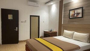 A hotel room with a bed, a desk, a chair, a nightstand, and a wall-mounted air conditioner.