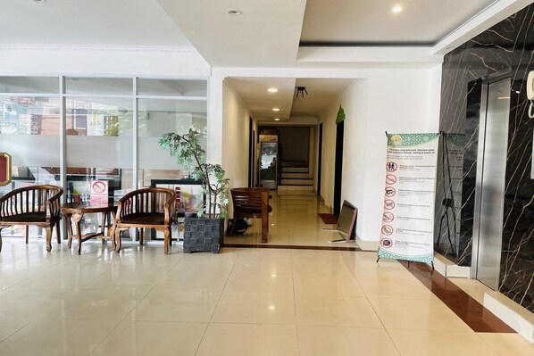 A hotel lobby with a reception desk, wooden chairs, a potted plant, and a marble wall.