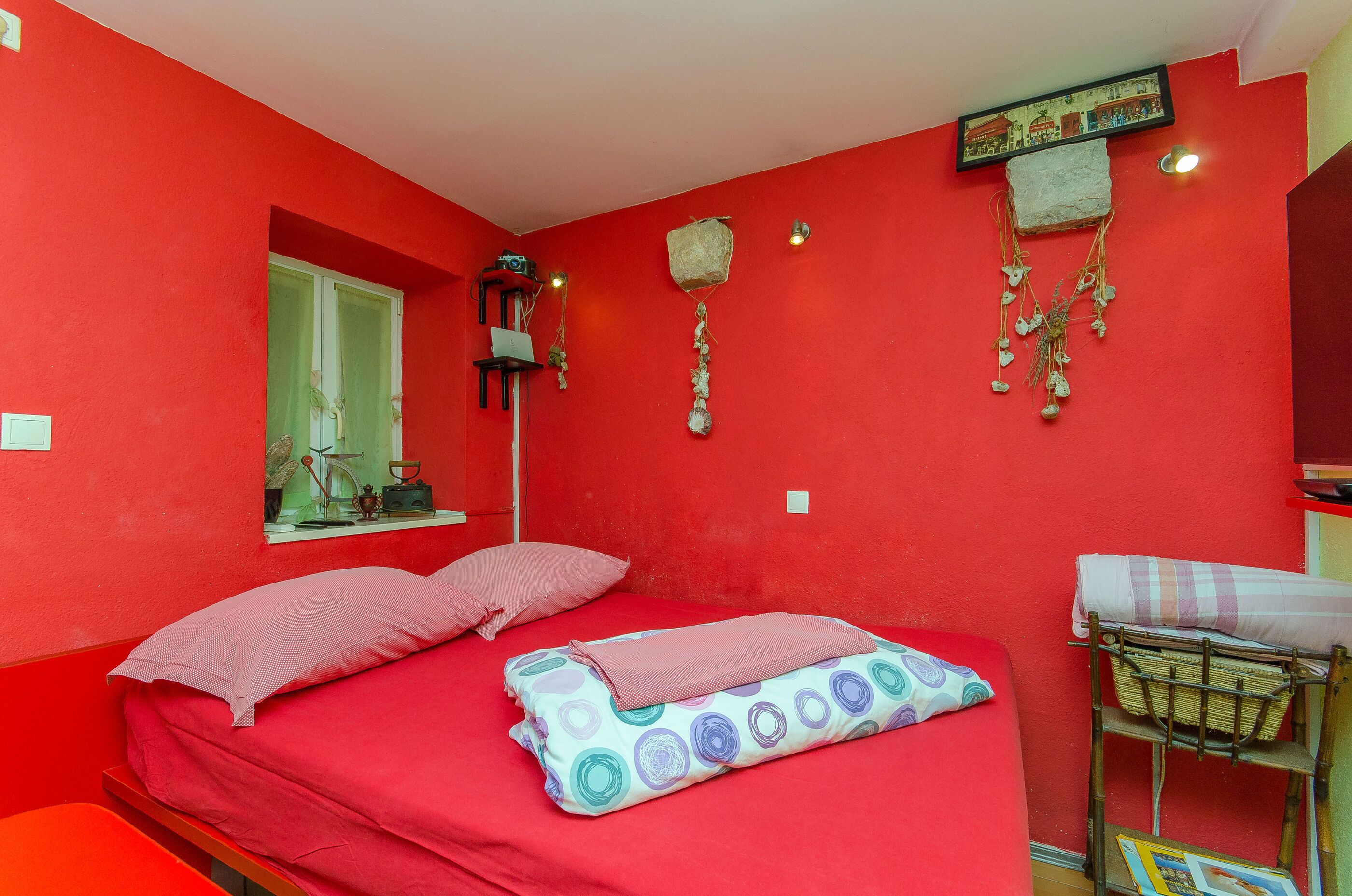 Une pièce aux murs rouges, avec un lit, des oreillers, une petite fenêtre, des décorations murales et une étagère sur laquelle sont posés divers objets.