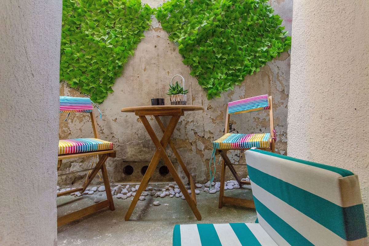 A small outdoor seating area with a round wooden table and two striped chairs, surrounded by a textured wall and greenery.