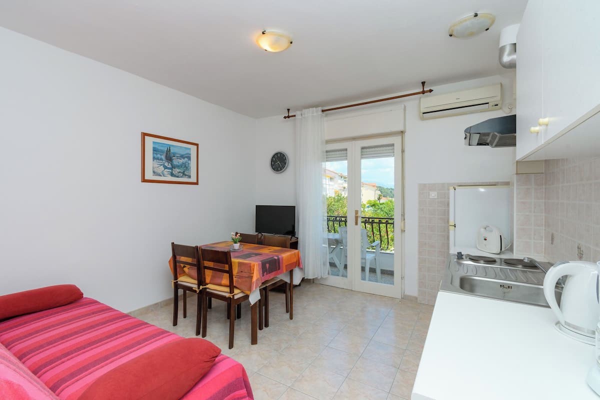 A hotel room with a bed, a dining table with chairs, a TV, an air conditioning unit, a kitchen area with a sink and stove, and a balcony with a view.