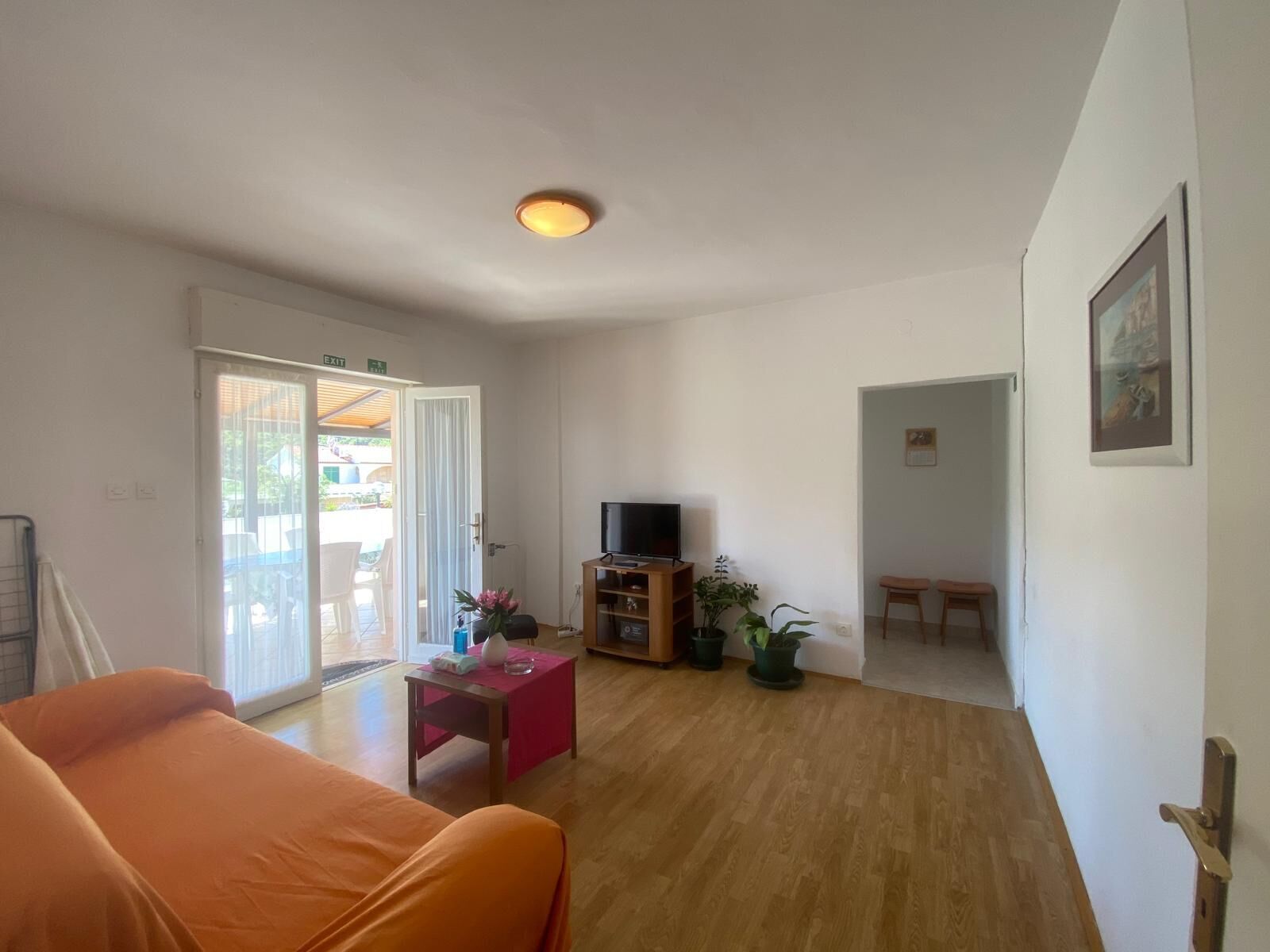 A living room with a sofa, a TV on a wooden stand, a small table with a vase, and a door leading to an outdoor area.