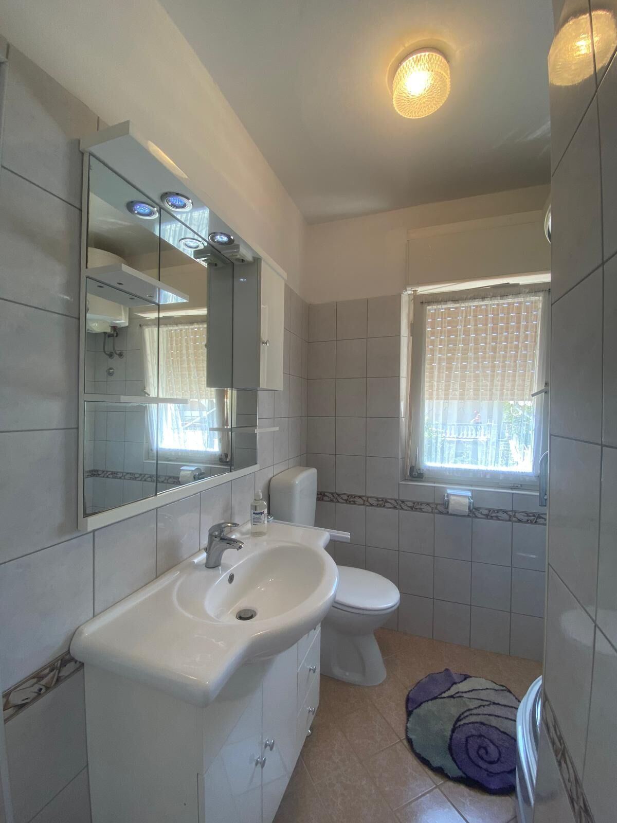Et badeværelse med en hvid vask, toilet og spejl. Der er et vindue med gardin og en lille måtte på gulvet.