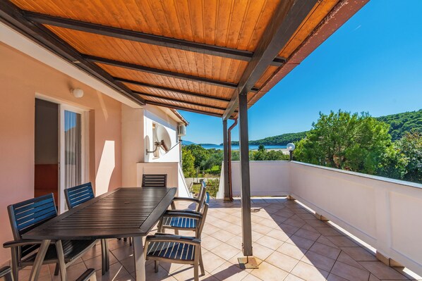 Un espacio de estar al aire libre techado con mesa y sillas, una puerta, y vistas al mar y las colinas.