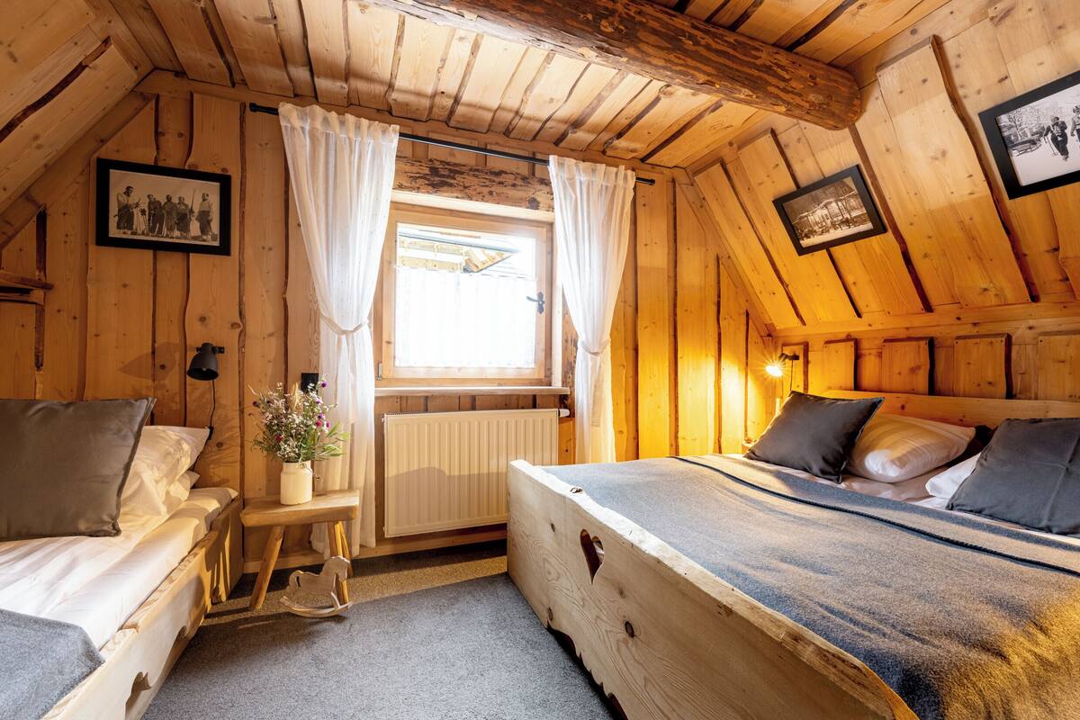 A wooden cabin bedroom with a bed, a small table, a radiator, and a window with curtains.