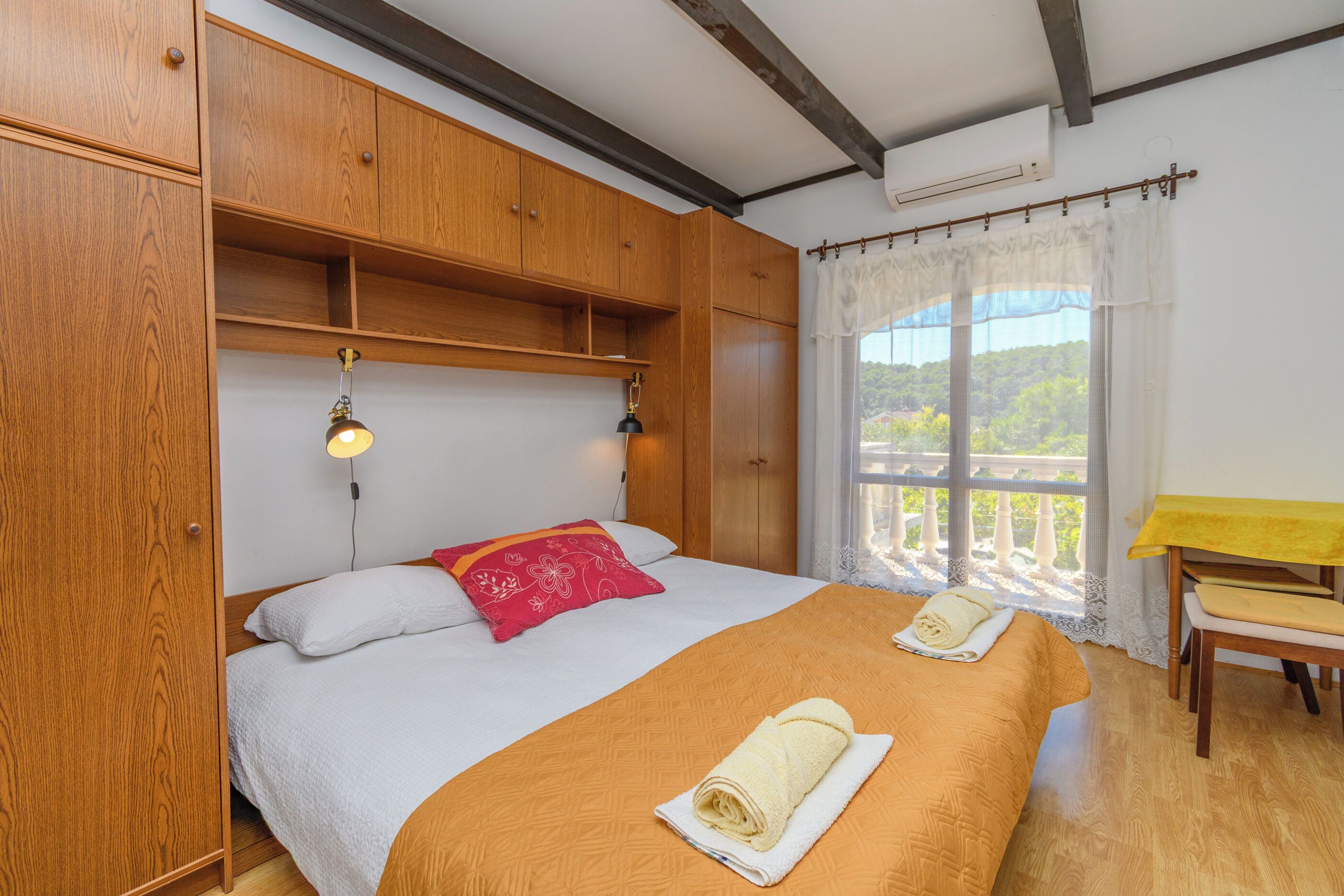 A bedroom with a bed, wooden wardrobe, bedside lamps, a window with a view, and a small table.