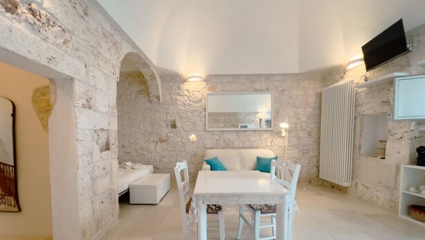 A room with a stone wall, a white sofa, a dining table with chairs, a mirror, and a TV mounted on the wall. - Suite Ninfea – Historic Home in the Heart of Ostuni (Ostuni)