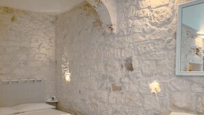 A room with a stone wall, a bed with white linens, a bedside table, and a wall-mounted light fixture. - Suite Ninfea – Historic Home in the Heart of Ostuni (Ostuni)