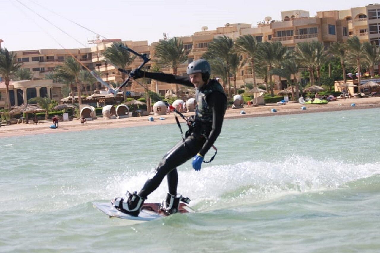 A person windsurfing on a calm body of water with a beach and palm trees in the background.