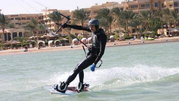 A person windsurfing on a calm body of water with a beach and palm trees in the background.
