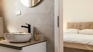 A modern bathroom with a white sink, a round mirror, and a beige headboard bed.