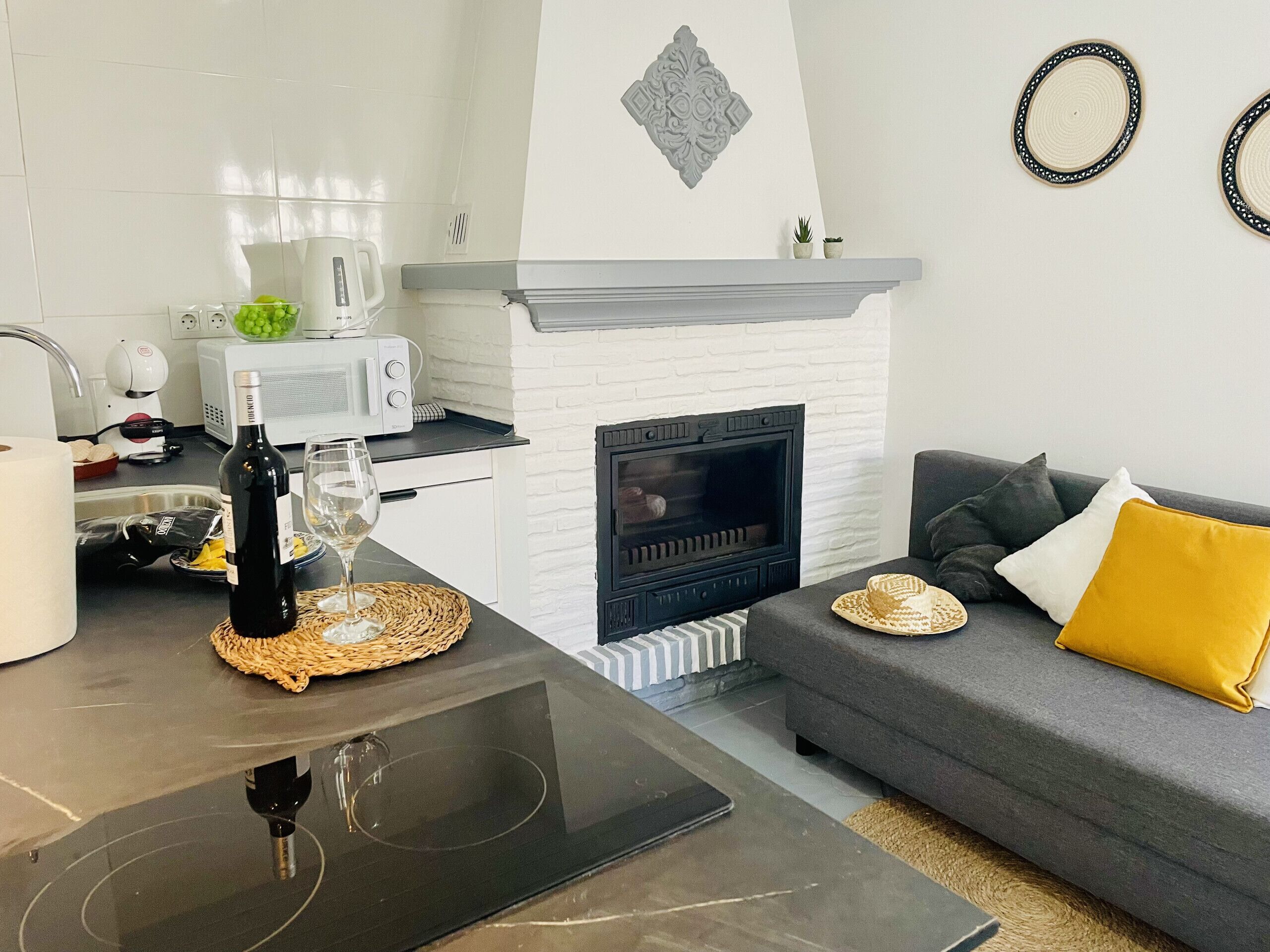 A modern kitchen with a microwave, toaster, and kettle, a dining table with a bottle of wine and glass, a fireplace, and a grey sofa with yellow cushions.