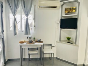 Una habitación pequeña y limpia, con una mesa, sillas, cafetera, televisor y estante con objetos decorativos.
