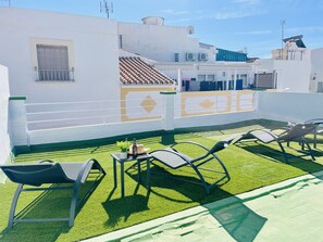 Terraza en la azotea con césped artificial, dos sillones de relax, una mesita con una botella y una planta, y un muro decorativo con patrones geométricos.