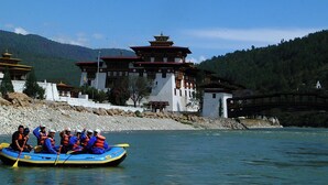 A group of people in a blue raft on a river, with traditional buildings and a bridge in the background.