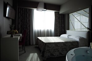 A hotel room with a bed, a desk with a fruit bowl, a chair, a large window with curtains, and a bathtub. - Moderno Hotel Roma (Rome)