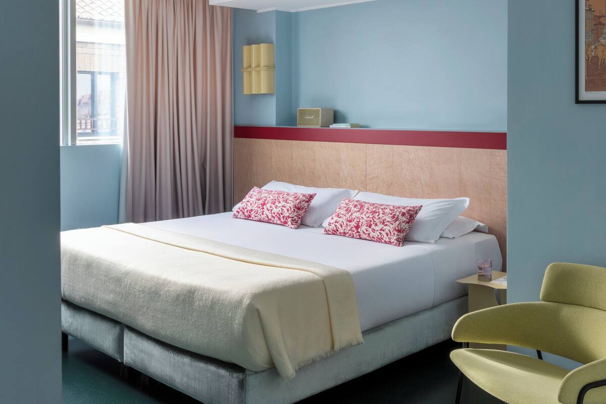 A hotel room with a bed, a chair, a bedside table, a lamp, and a window with curtains.