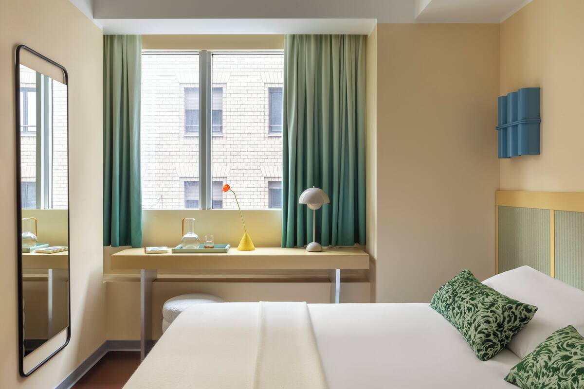 A hotel room with a bed, a desk with a lamp, a window with curtains, and a mirror.