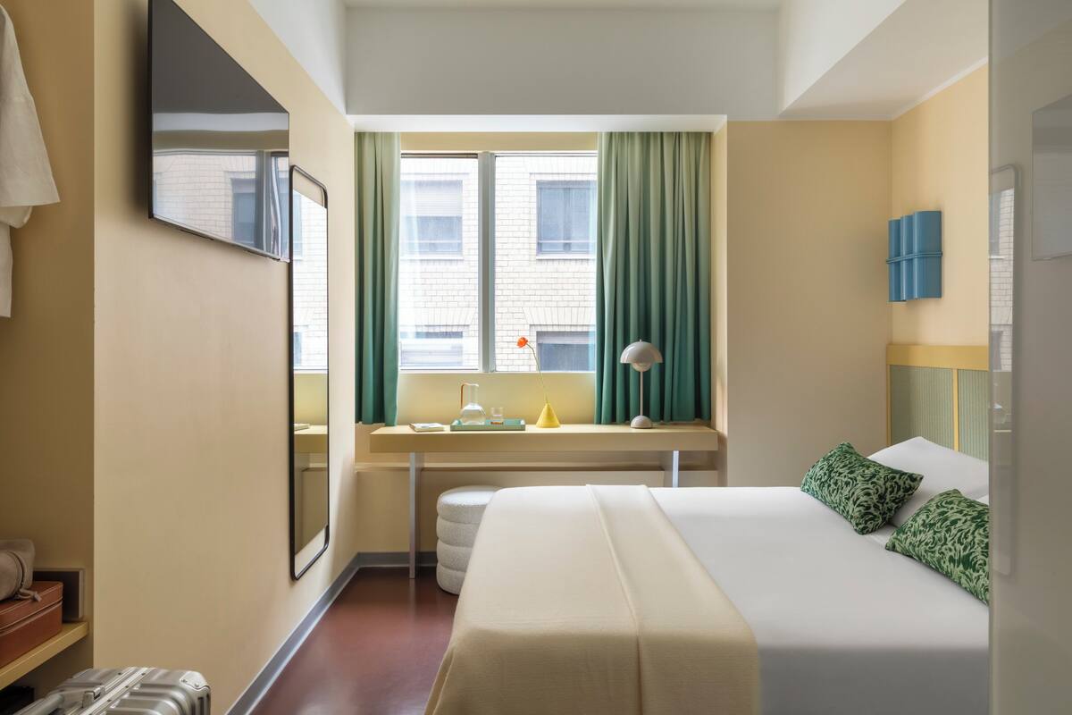 A hotel room with a bed, a desk, a window with curtains, a mirror, and a wall-mounted TV.