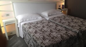 A hotel room with a bed, white headboard, two white pillows, a patterned bedspread, a nightstand with a lamp, and a small table with a laptop. - Moderno Hotel Roma (Rome)