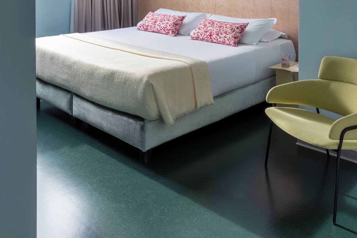 A hotel room with a bed, a chair, a bedside table, a lamp, and a window with curtains.