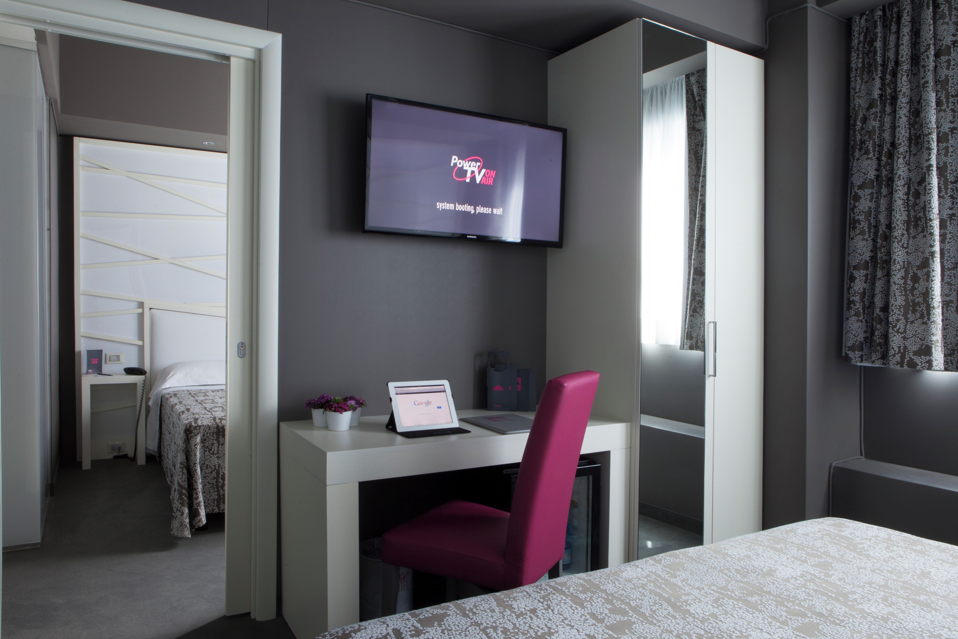 A modern hotel room with a bed, a desk with a laptop, a TV on the wall, and a chair.