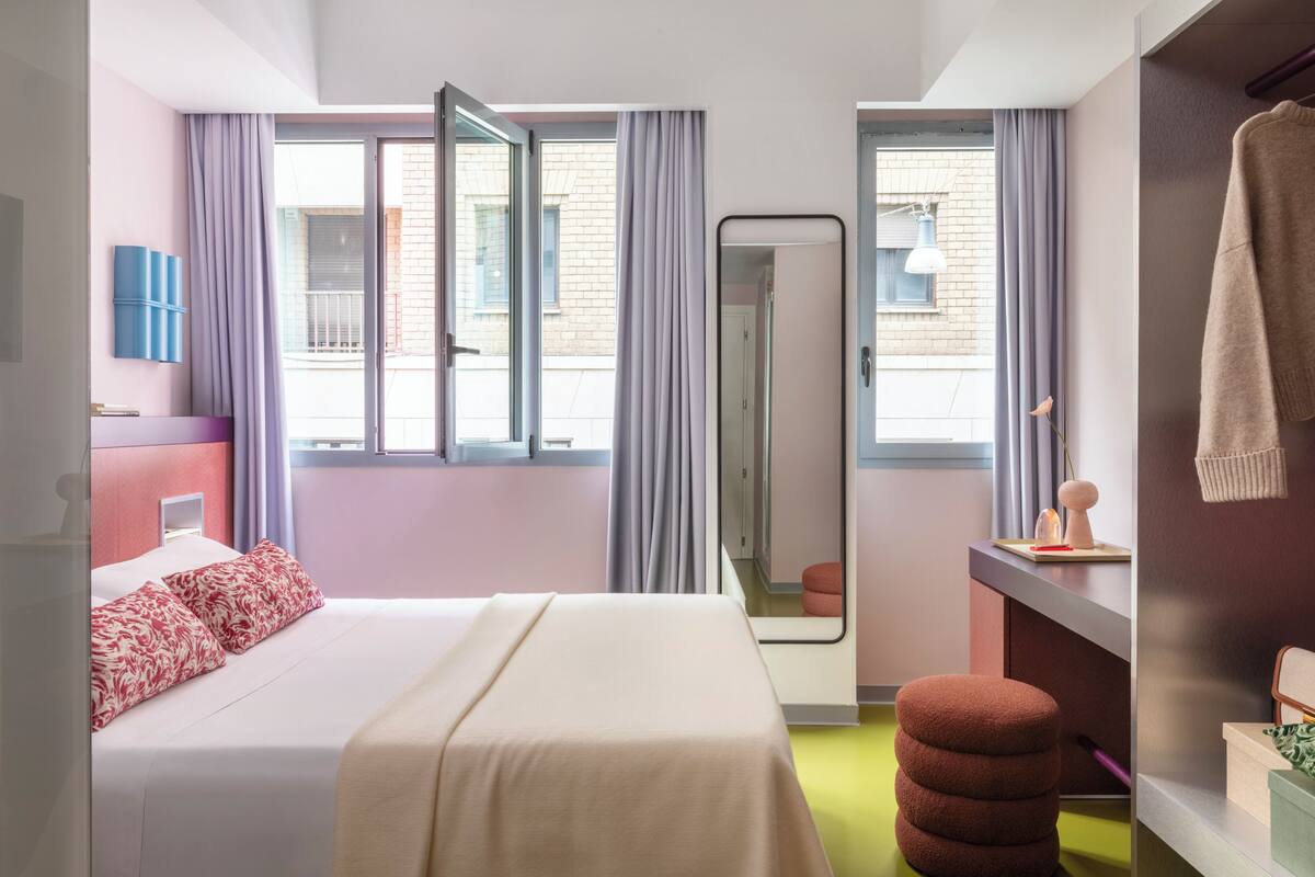 A hotel room with a bed, a mirror, a window with curtains, a small stool, and a towel rack.