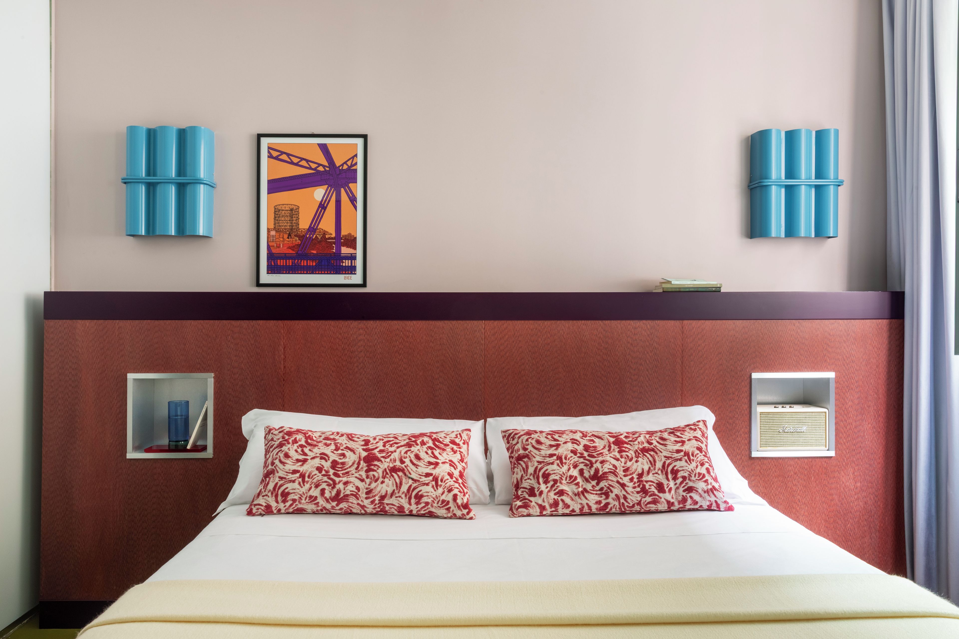 A hotel room with a large bed, red headboard, two pillows, a framed picture, two wall-mounted lamps, and a small shelf.