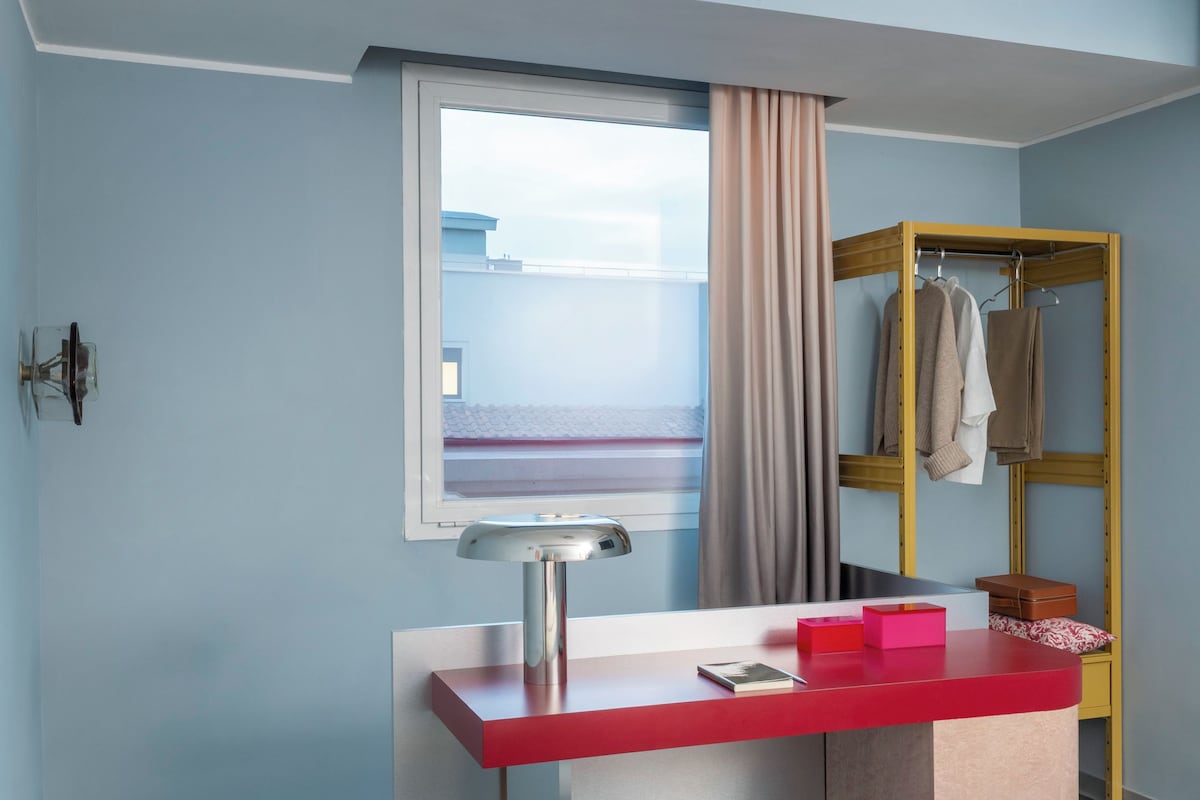 A compact hotel room with a bed, a desk with a red surface, a window with a view, a wooden wardrobe, and a small stool.