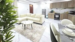A modern living room with a sectional sofa, a dining table set for two, and a kitchen area with wooden cabinets and a marble countertop.