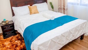 Ein ordentlich bezogenes Bett mit weißer und blauer Bettwäsche, ein Holz-Kopfbrett, ein kleiner Nachttisch mit einem Buch und ein flauschiger Teppich.