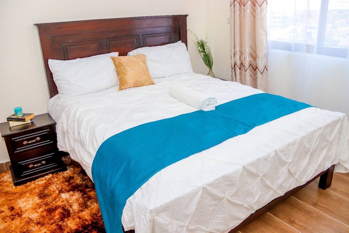 A neatly made bed with white and blue bedding, a wooden headboard, a small bedside table with a book, and a fluffy rug.