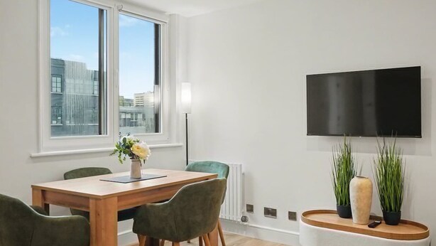 A modern dining area with a wooden table, green chairs, a vase with flowers, a flat-screen TV mounted on the wall, and a view of buildings outside. - Peaceful 1 bed apt in the Heart of Fitzrovia (London)