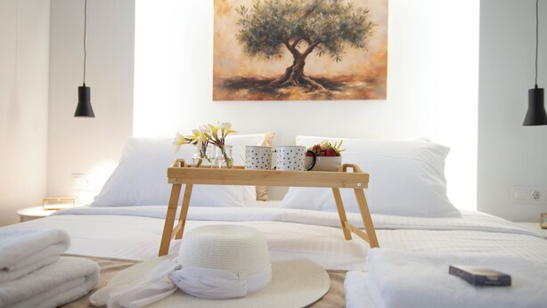 A neatly made bed with white linens, a bedside table with a tray of toiletries, a hat, and towels. - Casa Nefeli (Heraklion)