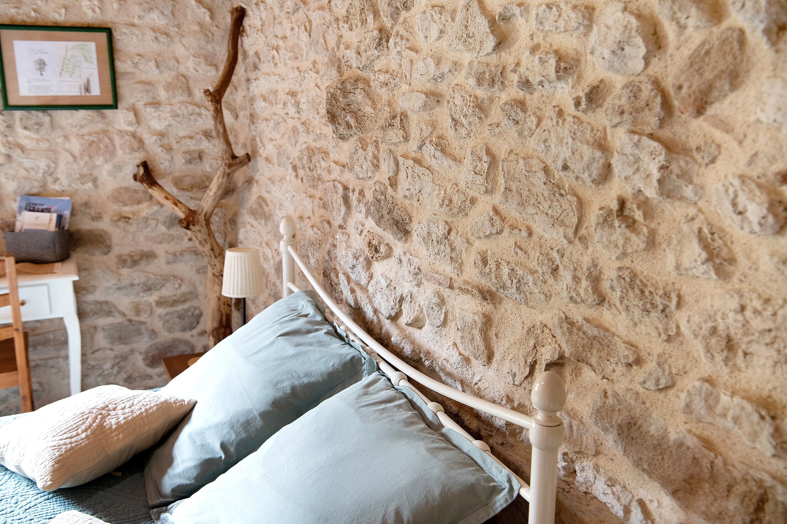 Una cama con cabecera de madera frente a una pared de piedra, una mesita de noche con lámpara y un cuadro enmarcado en la pared.