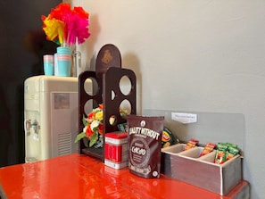 A table with a water dispenser, a coffee machine, a flower arrangement, a snack container, and a sign indicating a food and drink gift area.