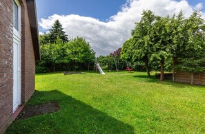 A backyard with a brick wall, a grassy lawn, trees, and a playground with a slide.