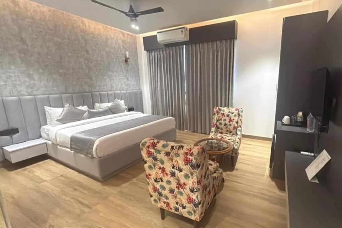 A modern hotel room with a bed, two chairs, a small table, a TV, and an air conditioning unit.
