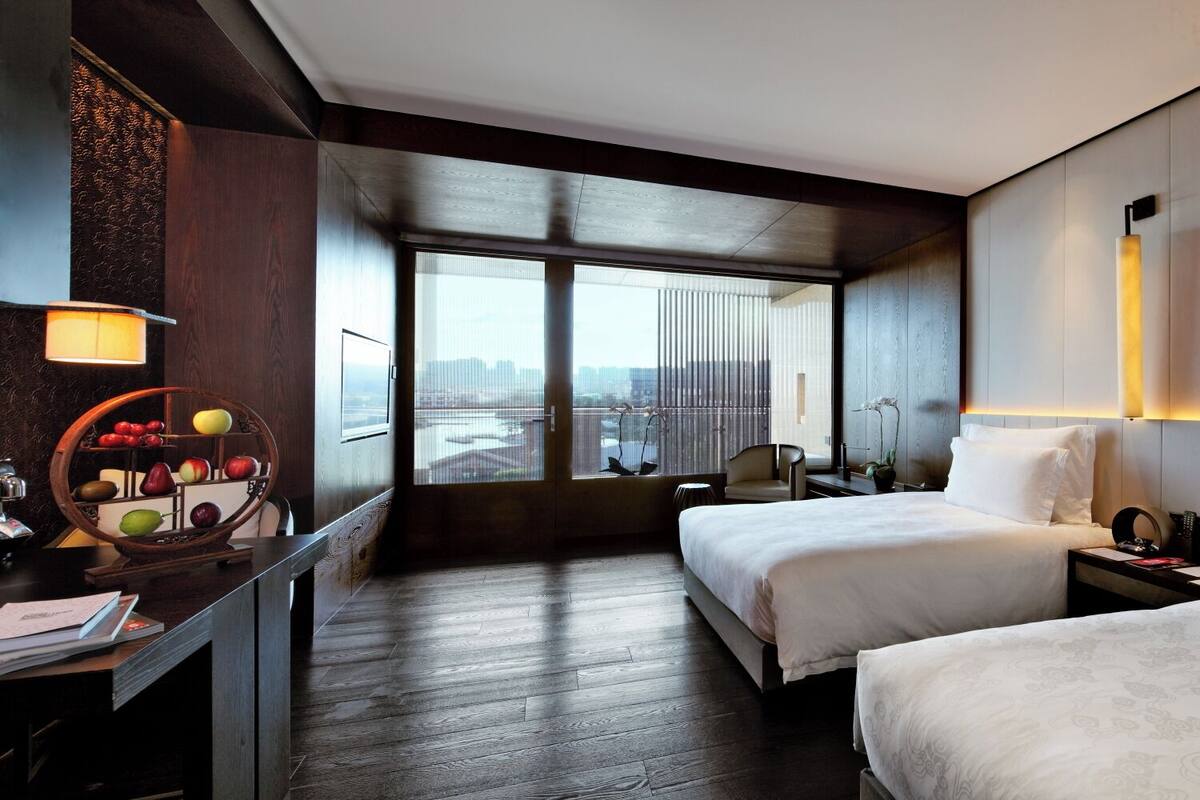 A hotel room with a bed, a desk with a lamp, a chair, and a view of the city.