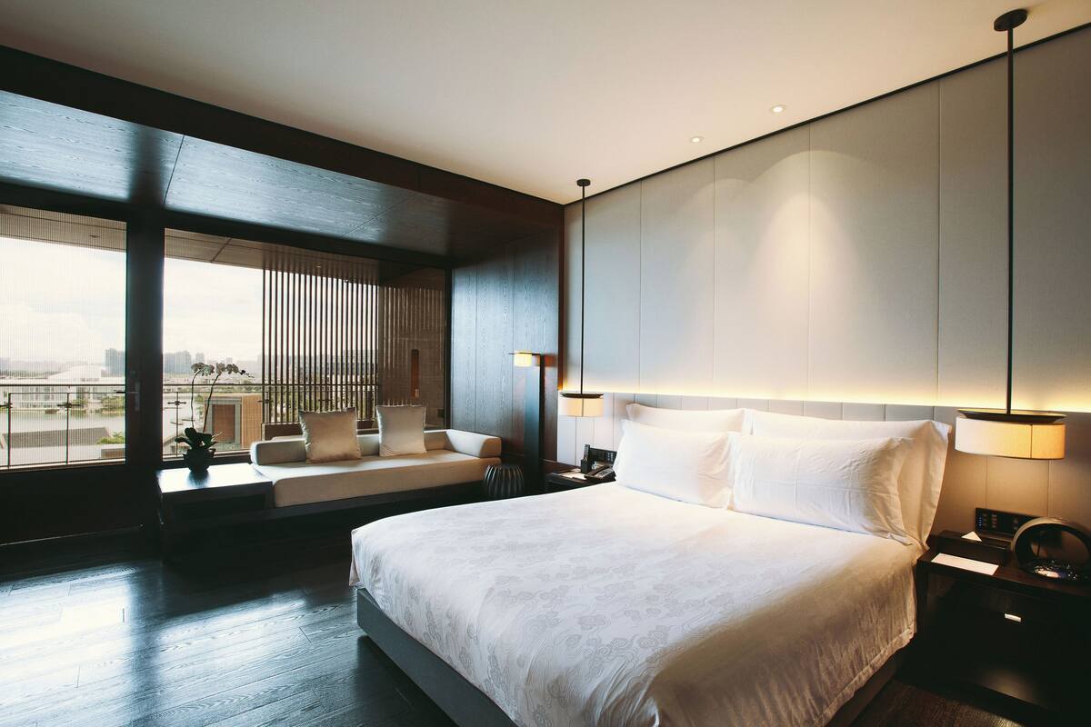 A modern hotel room with a large bed, a seating area with a sofa and chairs, a large window with a city view, and a wall-mounted lamp.
