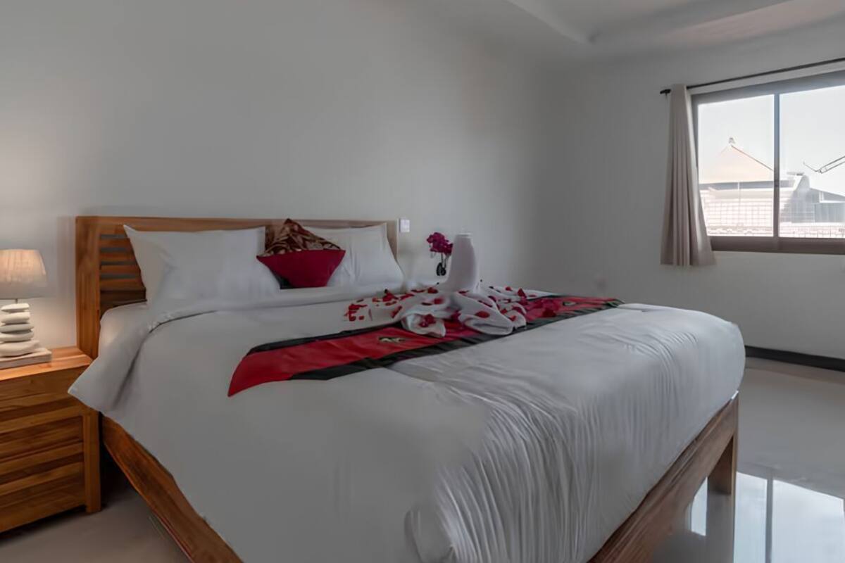 A neatly made bed with white bedding and red accents, a wooden headboard, a bedside lamp, and a window with a view of buildings.