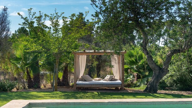 Una cama exterior junto a la piscina, con dosel y rodeada de exuberante vegetación.