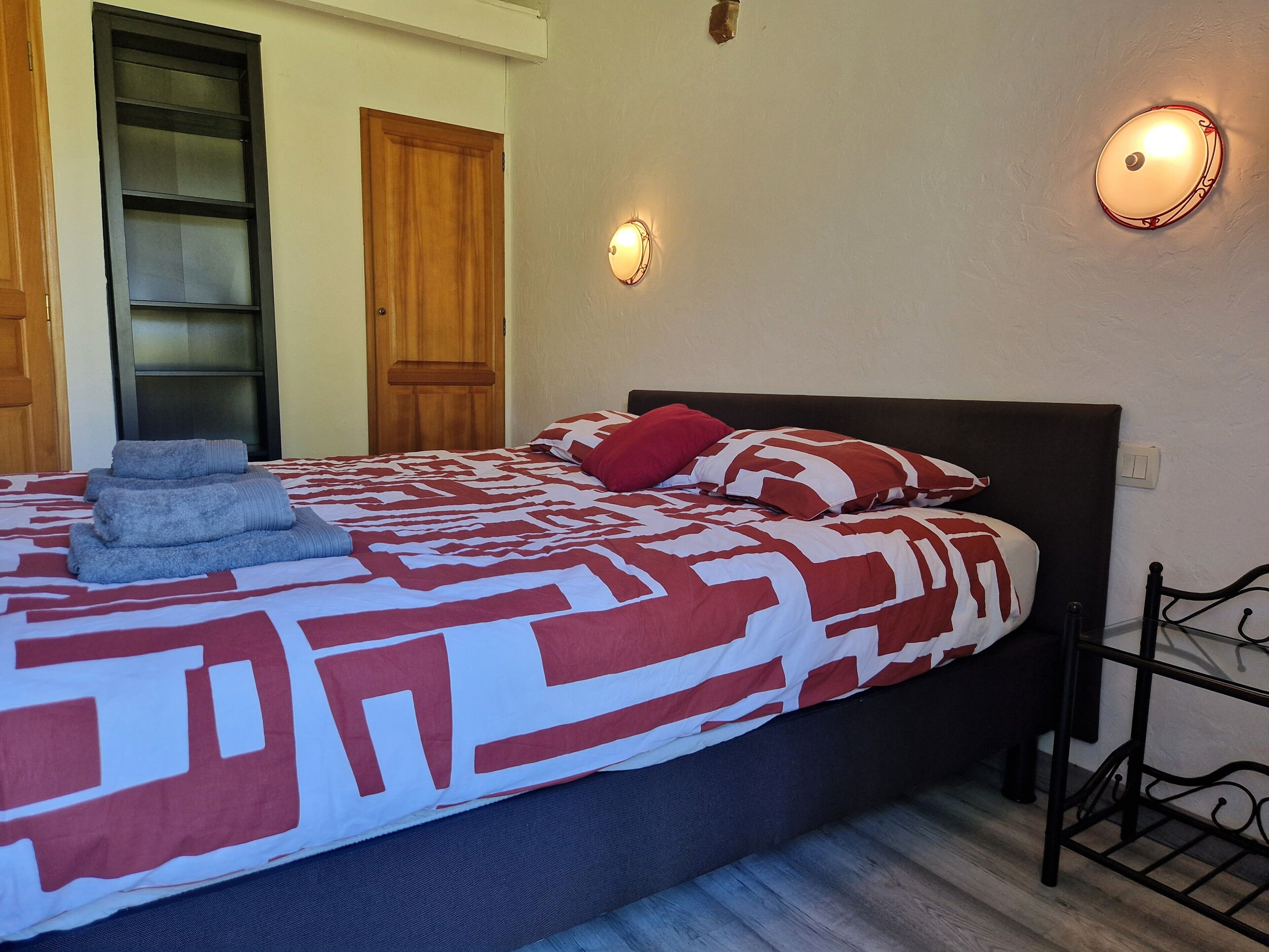 Een slaapkamer met een bed, nachtkastje, een lampje aan de muur en een deur.