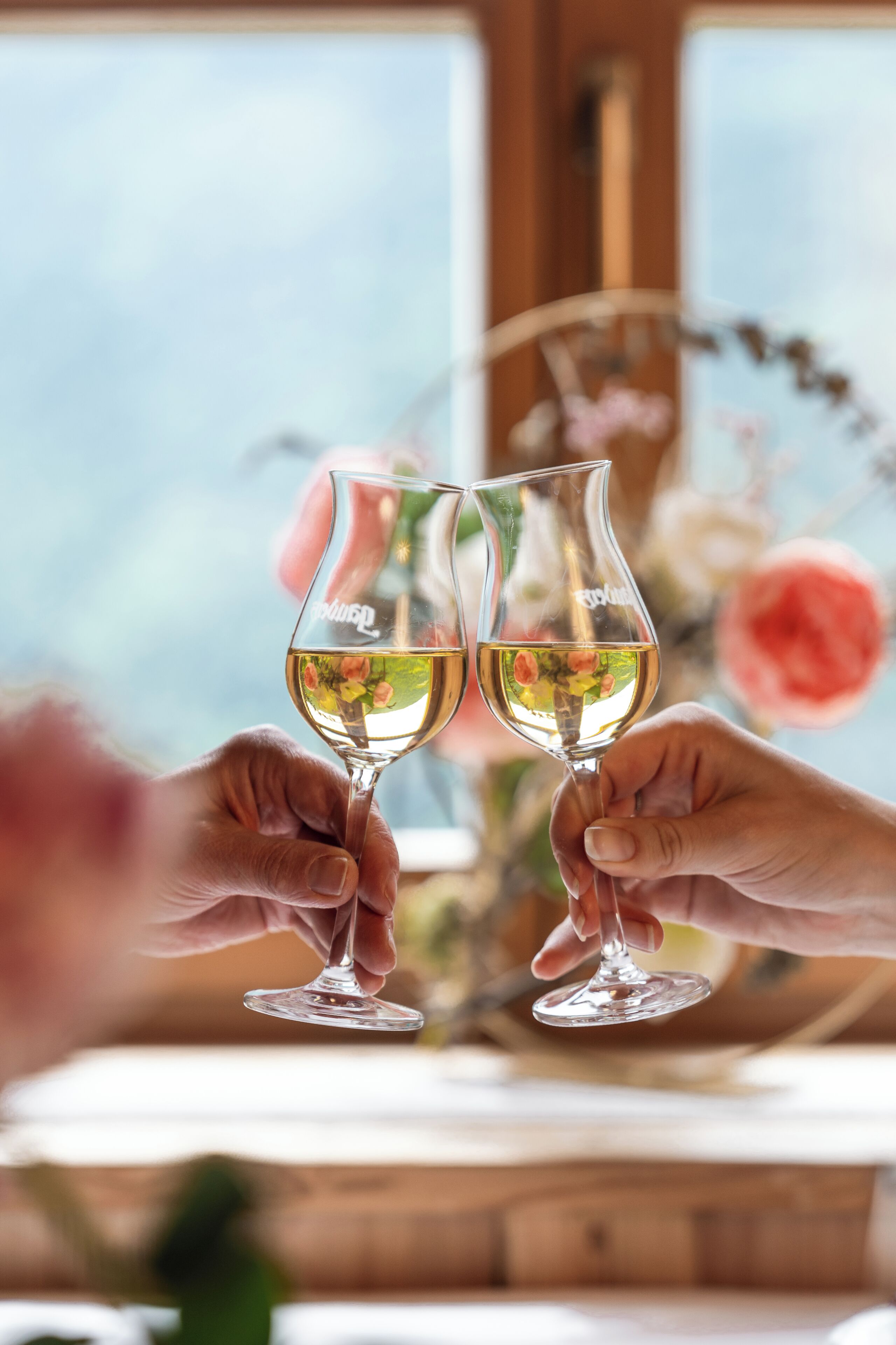 Two hands holding wine glasses with a floral arrangement in the background.