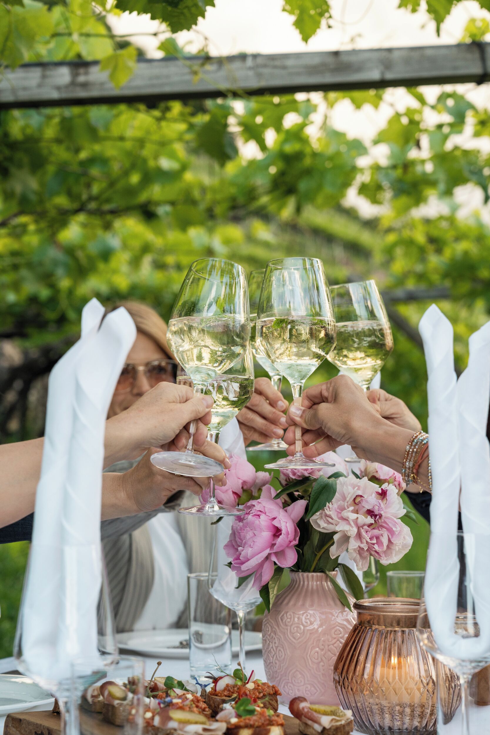 Un groupe de personnes trinquant avec des verres de vin autour d’une table en plein air, agrémentée d’un arrangement floral et d’un chandelier.