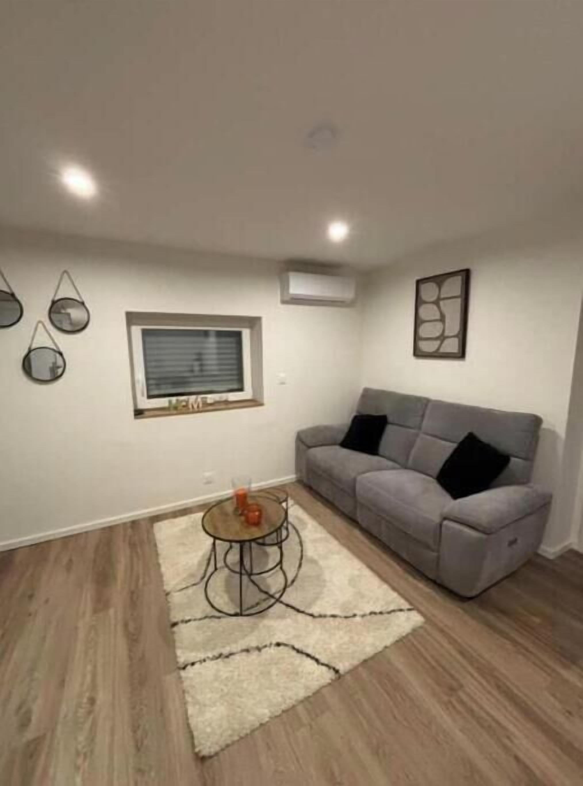A living room with a grey sofa, a small round table with a glass, a window with blinds, and wall-mounted air conditioning.